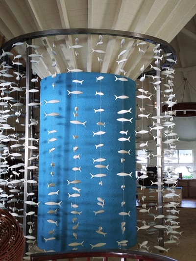 This is a medium shot, eye-level photograph of an interior space at the OBLU SELECT Sangeli resort on Gulhi Falhu, Maldives. The central focus is a large, cylindrical structure covered in a vibrant, textured teal-blue material. Suspended around this structure and hanging from a circular metal frame above are numerous white, flat, cut-out fish shapes. These fish are strung on thin wires at varying lengths, creating a visually dynamic mobile that cascades downwards. Some of the fish are oriented horizontally, while others are angled.
The ceiling above is constructed with exposed wooden beams and white paneled sections. The background to the right of the teal cylinder reveals a glimpse of the resort's interior, including wooden furniture, windows with natural light filtering through, and what appears to be cabinetry or shelving. In the foreground on the left, a portion of a dark brown woven wicker chair is visible. The overall impression is one of a decorative art installation within a resort setting. There are no people or animals visible in the frame. The lighting appears to be a mix of ambient and artificial.