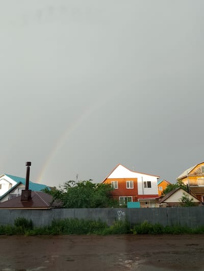 A residential area in Birsk, Russia, is captured under a gray, overcast sky, with a faint rainbow arching across the upper portion of the image. The foreground is dominated by a wet, muddy road, reflecting the dull light. A weathered, corrugated metal fence runs horizontally across the mid-ground, separating the road from the properties beyond. Behind the fence, a line of houses and trees is visible.
The houses exhibit a variety of architectural styles and colors. To the left, a house with a distinctive turquoise roof is partially visible. Adjacent to it is a house with a dark brown, gabled roof and a prominent brick chimney. Further to the right, a two-story house with white and reddish-brown brick lower levels and white upper levels stands out, its windows reflecting the muted light. Another smaller house with a beige facade and a brown roof is nestled behind it. To the far right, a wooden house with a yellow-orange facade and a steeply pitched roof is visible. Lush green trees and foliage are interspersed among the houses, indicating a well-established residential neighborhood. A faint chalk drawing of a heart with a swirl is visible on the fence. The overall scene suggests a post-rain environment, with the dampness of the road and the presence of the rainbow pointing to recent precipitation.