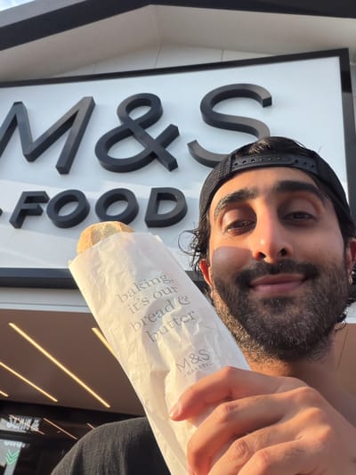 A man is taking a selfie outside an M&S Food store. He is wearing a black baseball cap and a dark shirt. He is holding a paper bag with a loaf of bread inside. The M&S logo and "FOOD" are visible on the store. The lighting suggests it is late afternoon or early evening. The specific location is the M&S Food store in Cannock Chase, United Kingdom.