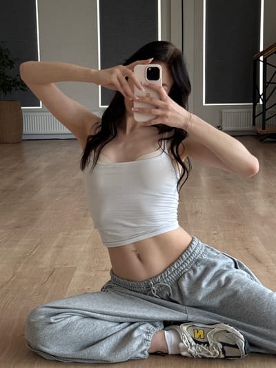 This media file shows a person taking a mirror selfie in a room with light wood flooring. The person is lying down, with their legs bent and one foot resting on the other. They are wearing a white tank top, grey sweatpants, and athletic shoes. The tank top exposes their midriff, revealing a belly button piercing. The room appears to be a home, with a white vent visible on the wall in the background, and a glimpse of furniture. The lighting suggests it is daytime. The image is an indoor shot, and there is no visible text.