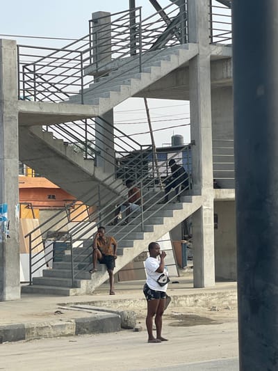 A concrete overpass with metal railings dominates the view. Three individuals occupy the stairs. One person is on a landing, another on a lower step, both appearing to be looking at a phone. A third stands on the sidewalk adjacent to the structure. The entire image is angled as if captured from an oblique vantage point. Overhead, a dark cylindrical object is partially visible.