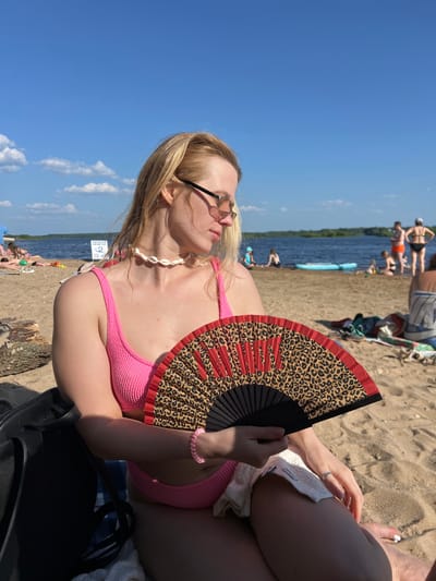 In Ust'ye, Russia, a woman with blonde hair and glasses sits on a sandy beach, holding a leopard-print fan. She wears a pink bikini and a seashell necklace. In the background, a body of water is visible with several people scattered along the shore and in the water. The sky is blue with scattered clouds.