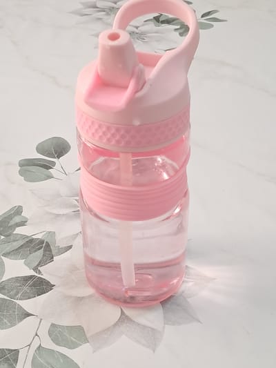 A pink plastic and clear plastic water bottle with a straw is standing upright on a white surface with a floral pattern. The bottle is partially filled with a clear liquid, and has a pink lid with a pink straw extending from it. The lid has a handle and a flip-up spout. The bottle is approximately 6 inches tall. The background surface is white and features a faint, grey, floral design with leaves and petals. The lighting is bright, casting subtle shadows. No people are present in the image.
