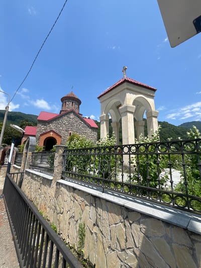 A medium shot captures a stone church with a red tiled roof in Adjaristskali, Georgia. To the right of the church is a light-colored, open-air bell tower with a red tiled roof and a cross at its apex. Both the church and bell tower are partially obscured by a black metal fence and lush green foliage. The church is constructed of rough-hewn stones, with a prominent arched entranceway. The bell tower features four arched openings supported by stone pillars. Behind the structures, a verdant, tree-covered mountainside rises under a clear blue sky dotted with a few white clouds. A stone wall runs along the foreground, topped by the black metal fence. A narrow asphalt pathway or road is visible to the left, bordered by a black metal railing. A utility pole with a light fixture stands to the left of the church.