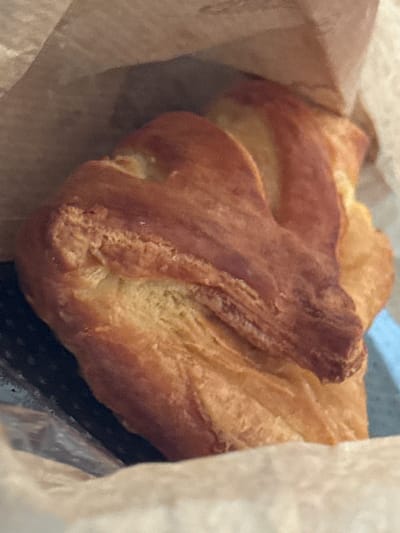 A close-up, eye-level shot captures a golden-brown croissant resting inside a brown paper bag. The croissant, which appears to be flaky and layered, is positioned in the center of the frame, taking up most of the space. Its surface exhibits a rich, baked texture with some darker brown hues indicating where it is more browned. The paper bag is visible surrounding the pastry, with its crinkled texture adding a sense of depth. The lighting is soft, casting gentle shadows and highlighting the croissant's golden color. The background is a blur of muted tones, suggesting an indoor setting, potentially a bakery or kitchen. There are no people visible in the image.