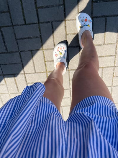 The media file shows a low-angle shot of a person's legs and feet, wearing white Crocs with colorful charms and a blue and white striped dress. The person is standing on a paved walkway made of rectangular gray bricks, with some green moss growing in the cracks between the bricks. Sunlight casts a strong shadow across the walkway. The scene is outdoors, with the bright sunlight and clear shadows suggesting it is daytime, likely a sunny day. The focus is on the legs and feet, with the dress flowing around the thighs. The overall impression is of a casual, warm-weather setting. The location is identified as Holíč, Slovakia. There is no visible text in the image.