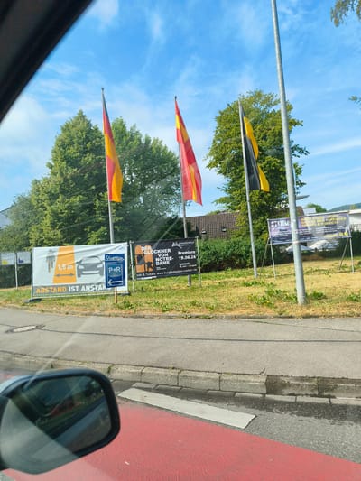 The image is a photograph taken from inside a vehicle, likely a car, looking out at a street scene in Lörrach, Germany, on a sunny day. Three German flags, a black, red, and yellow tricolor, are prominently displayed on flagpoles. In the background, there are trees, buildings, and a clear blue sky with some wispy clouds.

Several signs are visible along the roadside. One sign, featuring an orange background with a graphic of a cyclist and a car, displays "1,5 m" and "ABSTAND IST ANSTAND" (Distance is Decency), indicating a public health message. Another sign shows the logo "Stimmen Festival" and "P+R" with an arrow, suggesting a parking and public transport connection related to the festival. Further to the right, there is a poster advertising "DER GLÖCKNER VON NOTRE-DAME" (The Hunchback of Notre Dame) and "PREMIERE 19.06.26", along with details about the "BURGFFESTSPIELE RÖTTELN" (Rötteln Castle Festival).

The foreground shows the side mirror of the vehicle, reflecting the scene behind, and a red-painted area on the road, possibly a bicycle lane or a pedestrian crossing. The street is paved, with a concrete curb and some grass and weeds growing along the edge. The overall impression is a street scene on a day with good weather, possibly during an event or festival, with a focus on public information and safety measures.