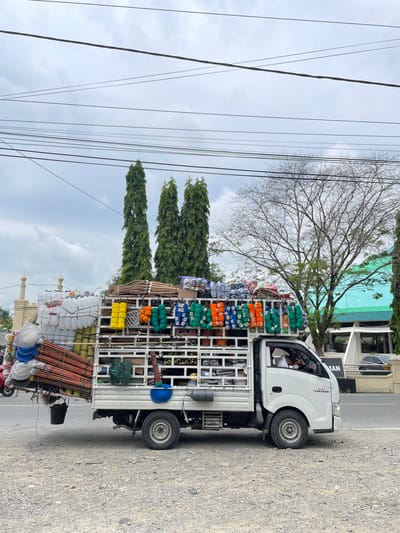 A side view of a white flatbed truck heavily laden with various goods is captured on a street in Pasar Paloh, Indonesia. The truck's bed is overflowing with items including bundles of long wooden poles, white plastic containers, colorful ropes or threads in shades of yellow, orange, and green, and what appear to be brooms or brushes. A blue bucket is hanging from the back of the truck. The driver, a person wearing a white shirt, is visible inside the cab, appearing to be resting their head on the steering wheel. In the background, several tall, slender trees line the street, along with a building with a green roof and white facade. Overhead, a tangle of power lines crisscrosses the overcast sky. The street surface is gravelly.