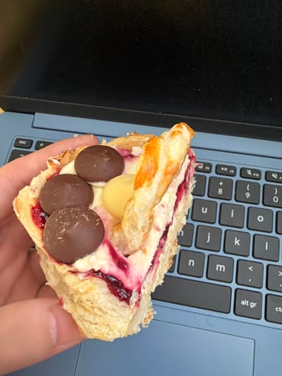A person's hand holds a partially eaten pastry, seemingly near or on a laptop keyboard with black keys and a blue body. The pastry has flaky layers, a filling of cream, red jam-like substance, and both brown and yellow sphere-shaped sweets or fillings. The background includes the laptop and a black surface. The image provides no information about the location within Pentre, United Kingdom, nor does it contain any information about specific actions, interactions, or people.