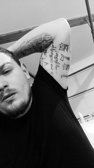 This black and white photograph depicts a male subject in an indoor setting within Senec, Slovakia. The subject is seen from the chest up, positioned at an angle with his left side closer to the camera. He has short hair and a beard/stubble on his visible left cheek and jawline. He wears a dark, possibly black, short-sleeved garment. His right arm is bent and raised, with the hand obscured, likely positioned behind his head or neck. The arm displays multiple tattoos: a script tattoo on the inner bicep reading "is one authority for- my father!", an elaborate, dark-shaded design further up the arm, and gothic-style lettering including "7" and other characters on the underside of the forearm/bicep. The background is light-toned, possibly a wall or ceiling. A dark, diagonal structural beam or frame is visible in the upper right, and a white, cylindrical bottle is present on a surface in the lower right background. The subject is looking directly towards the camera with a neutral expression.