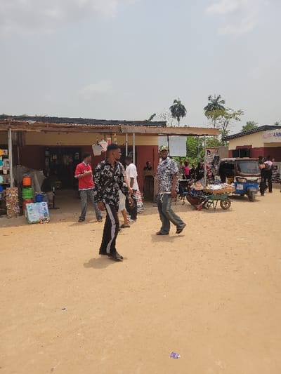 The scene depicts an unpaved, sandy open area functioning as a public space or market in Candelaria, Colombia, under a lightly overcast sky. The ground is a light brown, indicating dirt or sand, with scattered litter including a purple wrapper.

In the foreground, a male subject walks from left to right, wearing a black and white patterned long-sleeved shirt, black track pants with white stripes, and dark shoes. His shadow is cast to his left. Walking towards him from the right, another male subject wears a light-colored patterned short-sleeved shirt, dark pants, and a baseball cap. His shadow is cast to his right.

In the midground, to the left, a building with a dark red-brown base and a makeshift wooden and corrugated metal overhang is visible. Displayed outside this structure are stacked green cylindrical objects, various packaged goods, and a black standing fan. A male subject in a red short-sleeved shirt and patterned pants stands near this building. Another male subject in a white shirt is also present nearby.

Towards the right midground, a green, single-wheeled pushcart containing various items is positioned on the ground, seemingly attended by an unseen individual. Behind the cart, a light blue three-wheeled motorized vehicle is parked adjacent to a light-colored building. This building displays a partial sign reading "COLLECT A VOLT." Multiple individuals are present around these vehicles and buildings, including one person in a pink top and another in dark clothing on the far right. The background features scattered trees and foliage, indicating a vegetated environment.