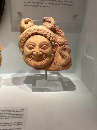 The image displays a terracotta architectural fragment, possibly a gorgoneion, at the Heraklion Archaeological Museum in Heraklion, Greece. The fragment depicts a grotesque, smiling face with wide eyes and elaborate hair or headdress. It is mounted on a clear acrylic stand against a plain, light-colored wall, creating a stark shadow on the surface behind it. Text labels are visible below and to the left of the artifact, indicating it is part of a museum exhibition. The lighting is even, illuminating the artifact and its display. The fragment has a weathered, earthy texture, characteristic of ancient terracotta. A number "4990" is faintly visible on the artifact's lower right side.