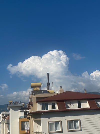 In Alanya, Turkey, a partially constructed building stands against a bright blue sky, dotted with large, fluffy white clouds. The building, seen from a slightly elevated angle, features a prominent, dark red tiled roof with dormer windows. The walls are painted in light colors, some with horizontal siding. On the rooftops of adjacent buildings, solar water heaters and satellite dishes are visible. A tall, dark chimney rises from one of the rooftops. The scene suggests a sunny day, possibly late morning or early afternoon, with a partly cloudy sky. The surrounding environment appears to be an urban or semi-urban area, with other buildings and distant hills or mountains visible in the background.