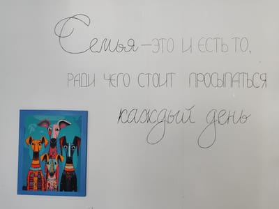 The image shows a wall with a handwritten quote and a framed artwork. The quote is in Russian and reads: "Семья — это то, ради чего стоит просыпаться каждый день" (Family is what it's worth waking up for every day). The artwork is a colorful, stylized depiction of four dogs. The background is a plain white wall. The image was taken in Snezhinsk, Russia.