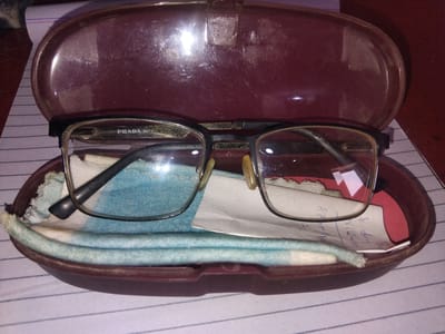 The image shows a pair of eyeglasses in a maroon-colored glasses case. The case is open and resting on a lined paper surface, likely a notebook or journal. Inside the case, the eyeglasses are positioned on top of a light blue cleaning cloth, and a folded piece of paper with writing is also visible. The eyeglasses have thin, metallic frames with what appear to be prescription lenses. The lenses show some smudges and dust. The inside of the glasses case is also lined with a soft material, and there are some scuff marks and scratches on the exterior of the case, indicating use. The lighting suggests an indoor setting, possibly a desk or table. The overall scene is a close-up view of personal belongings, with no indication of a specific location in Haveli Jhangranwali, Pakistan, or any people.