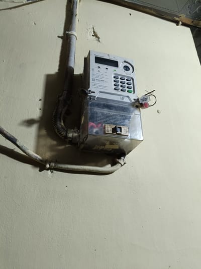 This image captures a close-up view of a smart electricity meter, specifically a prepaid meter manufactured by "PT. SMART METER INDONESIA" based in Tangerang, Indonesia, installed on a light-colored, textured wall in Cileungsi, Indonesia. The meter is predominantly grey and white, featuring a digital display panel at the top, a numerical keypad (0-9) below it, and various labels indicating specifications such as "3x220V," "50Hz," and a "GARANSI 5 TAHUN" (5 Year Warranty) sticker. A white corrugated electrical conduit descends from the top left, connecting to the meter's upper section, while a painted, rigid conduit or pipe runs horizontally beneath the meter.
Below the main meter unit, a clear, possibly plastic or glass, enclosure with metallic edges is secured. This enclosure reveals internal wiring, including red and white electrical cables, and what appear to be circuit breakers or fuses. A red tamper-evident seal or utility tag, bearing the inscriptions "23," "PZTL.IL 090600," and "DUB SMP1," is affixed to the right side of the meter assembly, securing the clear bottom enclosure. The wall itself is a uniform, pale cream or off-white color, exhibiting a slightly rough texture and minor surface imperfections. Strong shadows are cast on the wall from the vertical conduit, indicating a light source from the left. In the upper right corner, a section of a ceiling or upper wall structure shows some wear and discoloration. No individuals or active interactions are visible in the frame.