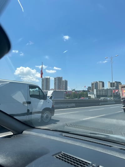 The image captures a daytime scene on a highway in Bayrampaşa, Turkey, likely during rush hour traffic. The viewpoint is from inside a vehicle, looking out through the windshield, which is partially obscured by the dashboard and a reflection of the sky.
Dominating the left side of the frame is a white panel van, its side facing the camera. The van appears to be in motion, suggested by the slight blur. The highway itself stretches across the middle ground, with multiple lanes indicated by white markings. In the distance, a cityscape rises, characterized by a cluster of modern, tall buildings. A prominent Turkish flag, red with a white crescent and star, is visible flying from a flagpole between two of the buildings.
Further in the background, other vehicles, including cars and what appears to be a truck, are visible on the highway and in the distance. A street light fixture with two lamps stands on the right side of the highway. The sky is a clear blue, with scattered white clouds and some thin streaks, possibly contrails. The overall impression is of a busy urban environment on a sunny day.