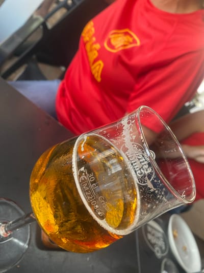 The image shows a close-up of a glass of beer, tilted to the right, with condensation on the outside. The beer is a golden amber color with a foamy head. The glass has some markings on it, including "30 cl" and "M2210866". In the background, a person wearing a red shirt with a yellow logo is visible. The setting appears to be outdoors at a cafe or bar, with a black table in the foreground. The presence of "San Pedro De Alcántara" in the prompt suggests this scene is located in the city of San Pedro De Alcántara, Spain.