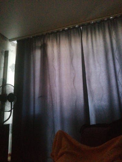 The image depicts an indoor scene, likely a room, dominated by dark gray curtains that cover a significant portion of the frame. The curtains are drawn, creating folds and shadows. To the left of the curtains, a portion of a white wall with a floral pattern is visible, along with the top of a black electric fan. The ceiling is visible in the upper part of the image. In the lower right corner, a blurry orange blanket or fabric is draped over an unseen object. The lighting appears dim, casting shadows and creating a somewhat obscured view of the environment. The location of Achinsk, Russia, is not discernible from the visual elements present in the image.