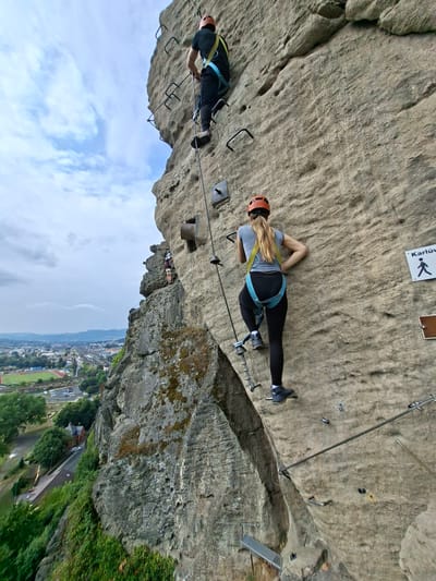 The image depicts a rocky cliff face with two individuals engaged in climbing, likely on a via ferrata route. The person in the foreground, a woman with blonde hair wearing a grey long-sleeved shirt, black leggings, a blue climbing harness, and an orange helmet, is ascending the rock face using metal rungs and cables. She is positioned mid-climb, with her right leg on a rung and her left hand gripping a cable. Above her, further up the cliff, a second person, a man wearing a black t-shirt, black pants, a yellow and blue climbing harness, and an orange helmet, is also climbing, his back to the camera.

The rock face itself is rough and textured, with various ledges and crevices. Metal fixtures, including rungs and cables, are bolted into the rock to facilitate climbing. A sign with a walking figure and text, partially obscured but potentially reading "Karlův," is visible on the right side of the rock face.

In the background, a panoramic view of the city of Děčín, Czechia, unfolds. The cityscape includes buildings, a railway track with a train, roads with cars, a stadium with a track, and a body of water. The sky is overcast with scattered clouds. The vantage point suggests the climbers are high above the city, offering a dramatic perspective. The overall scene captures an outdoor adventure activity set against an urban backdrop.