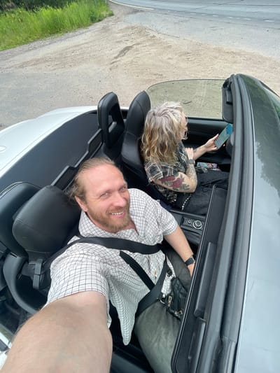 In Bolshaya Izhora, Russia, two individuals are seated in a convertible car. The person in the foreground, a male with blonde hair and a beard, is taking a selfie. He is wearing a checkered shirt and a seatbelt. To his right, a female with blonde hair is looking at her phone. She has tattoos on her arms and is wearing a plaid shirt. The car's interior is visible, with black leather seats and the convertible top down. The background shows a dirt road and green foliage, suggesting a rural or suburban setting. The lighting indicates it is daytime.