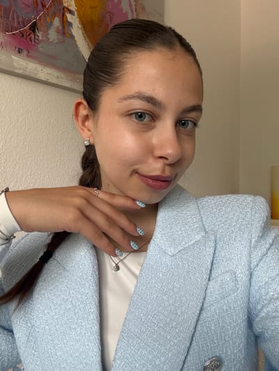 A woman is depicted in a close-up, tilted view. She has light skin, blue eyes, and her dark hair is pulled back into a braid. She wears a light blue tweed blazer over a white top, and a necklace with a pendant is visible. Her nails are painted light blue with black polka dots. She is wearing earrings and a ring. The background includes a partially visible painting and a light-colored wall. There is no information to place this image in Bruchköbel, Germany.