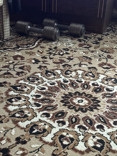 In Морозовка, Russia, two dumbbells are placed on a patterned rug next to a wooden piece of furniture. The dumbbells are metallic with layered weight plates. The rug features a complex design with floral motifs in shades of brown, beige, and black. The environment appears to be indoors, possibly a residential setting.