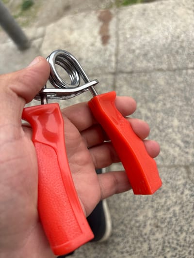 The image displays a human hand, exhibiting light skin pigmentation, holding a red and silver hand gripper. The hand gripper consists of two red, ergonomic, injection-molded plastic handles, one featuring a textured grip area, connected by a coiled chrome-plated steel spring and two steel pins. The device is held in a relaxed, open position. The background is out of focus, revealing a light grey paved surface, consistent with concrete or flagstones, marked by visible seams. A blurred, vertical metallic pole is discernible in the upper-left background, and a dark, indistinct object, possibly part of a shoe, is partially visible beneath the hand on the right. The scene is situated outdoors in the city of Lehvaz, Armenia.