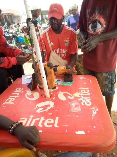 The scene depicts an outdoor commercial setting in Gana Daji, Nigeria, possibly a market stall. A dark-skinned male, centrally positioned, wears a red t-shirt with an Adidas logo, SL Benfica crest, and "Emirates Fly Better" text, along with a red "Airtel" baseball cap. He has a dark beard and is looking downwards, holding an orange electronic device in his right hand. A light-colored canvas bag hangs from his left shoulder.
He is seated at a red plastic table, visibly worn with white paint chipping, featuring "Airtel" branding in white text. A white pole, likely for a canopy or sign, protrudes through a central hole in the table. On the table, to his left, is a black mobile phone or device, and an orange power bank or rugged radio unit. To his right, several small clear plastic pouches, possibly containing SIM cards, are scattered alongside a green Nigerian Naira note and other currency. A document with "O Pay" visible is also on the table.
To the left, a person's hands in a red long-sleeved garment are visible, handling small items, possibly SIM cards. A dark-skinned hand with a multi-beaded bracelet rests on the table. To the right, partially visible, stands a person in a maroon t-shirt with a large eye graphic and dark-patterned shorts; their hands are clasped in front.
In the blurred background, several individuals are present, including a woman with a white head covering carrying a baby on her back and another woman in a blue top. Vehicles, possibly motorcycles, are faintly visible further back, suggesting a busy public thoroughfare. The lighting indicates daylight conditions.