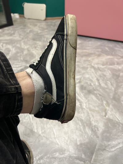The image shows a close-up, side-view of a black and white Vans sneaker on a person's foot. The sneaker is worn, with scuff marks and dirt on the sole and sides. The white stripe on the side is slightly discolored. The white sock is visible above the shoe and has a tear near the ankle. The person is wearing dark, possibly denim, pants. The background appears to be an indoor setting with a floor that has a marble-like pattern. In the background, there are blurry objects that suggest furniture or appliances, and a wall painted in dark green and a lighter pinkish-red. The image does not provide any information about the location in the city of Ленинский, Russia.