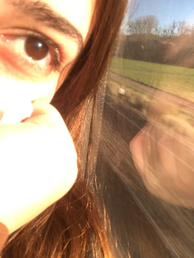 The scene depicts a close-up of a human subject observed from inside a vehicle, presumably in motion near Schwindratzheim, France. The left side of the frame features the right eye of an individual with a brown iris and pupil, and a portion of a dark eyebrow. Reddish-brown hair is prominent, extending from the left to the center of the image. A light-skinned hand or finger is visible below the eye. Direct sunlight illuminates the subject's face, hair, and hand. The right side of the image shows an exterior landscape viewed through a window, consisting of a green field, a light-colored dirt path, and bare deciduous trees under a clear blue sky. This external view is slightly blurred, consistent with travel. Overlaid on the window pane is a bright, streaked reflection of a human hand, distorted by light and motion.