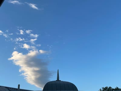 This image captures a clear blue sky with scattered white clouds. A large, elongated cloud formation dominates the left side of the frame, appearing wispy and textured. The lower portion of the image features the dark, curved roof of a dome-like structure, topped with a pointed spire. A dark, sloping roofline is visible on the far left, and a sliver of green foliage from a tree can be seen on the right. The lighting suggests it is daytime, likely late afternoon or early evening, given the clear sky and the presence of some dramatic cloud formations. There is no visible text in the image. The overall mood is serene and expansive, with the vast sky taking center stage.