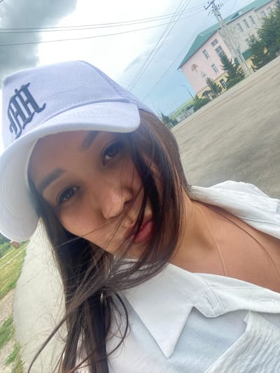 The image is a close-up selfie of a woman wearing a white baseball cap with a dark logo and a white collared shirt. She has long, dark hair and is looking at the camera. Behind her, there is a road, some grass, and buildings. The sky is cloudy. The setting appears to be an outdoor urban or suburban environment.