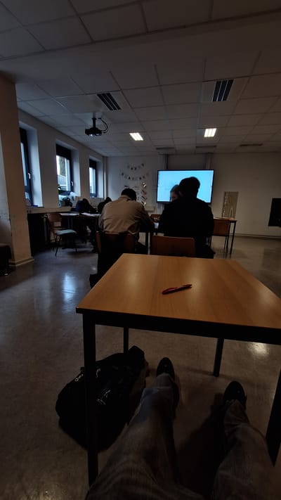 The image depicts the interior of a classroom or training room located in Bad Bergzabern, Germany. The room is dimly lit, with natural light entering through several tall, narrow windows on the left wall, and some overhead fluorescent light panels active in the suspended ceiling. A black projector is centrally mounted on the ceiling.

In the foreground, the legs of an individual wearing blue jeans and dark shoes are visible, extending under a wooden table. A black backpack or bag rests on the floor next to the individual's left leg. On the wooden tabletop directly in front of the camera, a single red pen is present.

In the midground, three individuals are seated at tables facing a large illuminated screen on the front wall. One person, wearing a light-colored collared shirt, is seated at a table on the left. A second person, dressed in a dark jacket or shirt, is seated more centrally, directly in front of the screen. A third individual with fair hair is partially visible to the right of the dark-jacketed person. The large screen displays text, partially legible as "E-Ma..." and "game...".

The back wall features a "HAPPY BIRTHDAY" banner composed of individual letter flags, along with decorative metallic stars. A long, dark counter or cabinet runs along the left wall beneath the windows. The floor is a light grey, possibly linoleum or concrete.