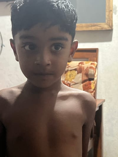An indoor, close-up portrait shows a young boy, likely a child or pre-teen, looking directly at the camera. The image is oriented sideways, with the boy's head positioned on the left side of the frame and his bare shoulders and torso extending to the right. He has short, dark, curly hair and dark, wide-set eyes, with a neutral or slightly pensive expression. He appears to be shirtless, revealing his medium-to-dark skin tone.

The background elements suggest a residential setting, possibly a home in Ulla, Sri Lanka. To the upper left of the boy's head, a portion of a wooden-framed mirror or picture frame is visible against a light-colored, textured wall. Behind his right shoulder and upper body, there is a section of wooden furniture, possibly a bed frame or headboard, with a patterned cushion or fabric featuring warm tones like orange, brown, and yellow. The lighting is soft and ambient, illuminating the boy's features. There is no discernible activity other than the boy posing for the photograph. No text is visible in the image.
