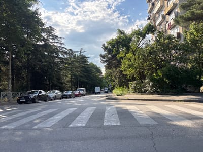 This is a street-level, eye-level, medium-wide shot of a pedestrian crossing in Tbilisi, Georgia. The scene is captured on a sunny day with a partly cloudy sky. Several cars are parked along the side of the road, and others are driving on the street. To the right, a multi-story apartment building is visible, with laundry hanging from the balconies. Lush green trees line the left side of the road, and more trees are present on the right side, behind a sidewalk. The pedestrian crossing is clearly marked with white stripes on the asphalt. The overall impression is a typical urban street scene with elements of residential life and traffic.