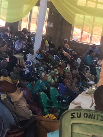 The image depicts a gathering of people, primarily adults, seated in rows of plastic chairs within a room in Nteje, Nigeria. The room is illuminated by natural light filtering through windows, some of which are draped with yellow fabric. There is white fabric draped overhead. People are dressed in a variety of clothing styles and patterns. Some chairs are blue or green. A container with the lettering "S.U UNITS" is visible.