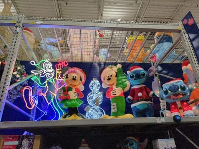 The image shows a shelf in a store displaying Christmas decorations. Mickey and Minnie Mouse figures, some holding Christmas trees, stand next to a light-up Goofy figure. Two Stitch figures, both wearing Santa outfits, are also on display. The shelves above are filled with various holiday-themed inflatables and light-up decorations. Price tags are visible on some of the items. The overall scene suggests a holiday shopping environment.