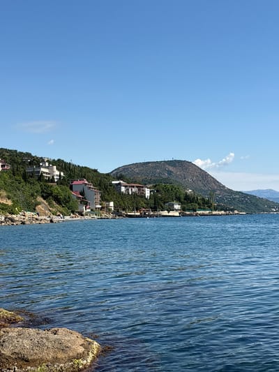 The image shows a coastal scene with buildings nestled on a hillside overlooking a body of water. The sky is a clear, bright blue with a few wispy clouds. The water is calm and a deep blue color, with gentle ripples on its surface. In the foreground, a large rock is partially submerged in the water.
The hillside is covered in lush green trees and vegetation, and several buildings of various sizes and architectural styles are visible. Some appear to be hotels or residential buildings, with white and beige facades, and red-tiled roofs. A paved walkway or road runs along the coastline in front of some of the buildings. A few scattered rocks and smaller boulders are visible along the shore.
In the distance, a large, rounded mountain dominates the horizon. The mountain is covered in sparse vegetation and has a few structures visible on its peak. The overall impression is of a peaceful, scenic coastal resort area. The location is identified as Lazurnoe, Ukraine.