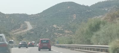 The image shows a highway in Guillena, Spain, with several cars driving on it. The road is surrounded by a mountainous, green landscape. In the foreground, a red car is visible, along with other cars on the highway. The sky is overcast.