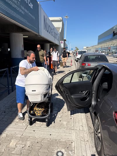 The image captures a moment in what appears to be Cinisi, Italy, specifically outside a car rental agency called "Nuova Sport Car." The scene is filled with bright daylight, indicating a sunny day. A man is standing near the open door of a dark gray or black car, holding a large, white baby carrier that sits in a stroller. He is wearing a white t-shirt and blue shorts with black and white sneakers.

Behind him and to the left, there are several other people walking. These individuals include a bald man in sunglasses and an olive-green shirt, a woman with dark hair, a man in a white shirt and khaki shorts, and another person with a suitcase. They seem to be associated with the same group.

The car rental agency's building is to the left, with its signage visible. Other parked cars and vans can be seen in the background, along with pedestrians walking further down the street. The ground is paved with square concrete blocks. In the distance, there's a glimpse of what might be the airport or associated facilities, including a tall pole with aviation-related equipment on top.