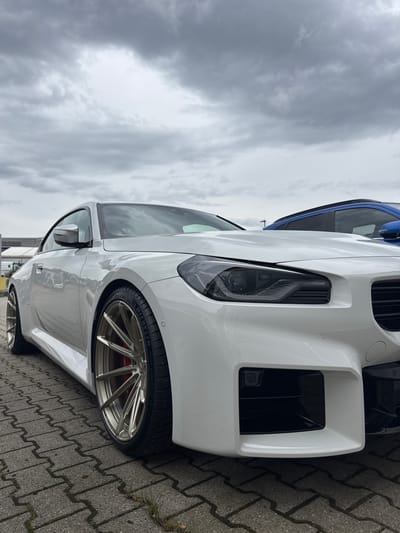 The image shows the front passenger side of a white BMW M2 CS. The car is parked on a grey, interlocking brick driveway. The sky is overcast, suggesting a cloudy or rainy day. In the background, a blue car is visible, also parked. The BMW has a large, aggressive front bumper with prominent air intakes. The headlight is visible, featuring a modern LED design. The car is equipped with a gold, multi-spoke wheel and a Michelin Pilot Sport 4S tire. The brake caliper is red. The BMW logo is visible on the wheel. The scene appears to be a car parking area, possibly in Wettstetten, Germany, given the location context. There is no discernible activity taking place, as the cars are stationary. The overall mood is neutral, with the focus on the car's design and details.