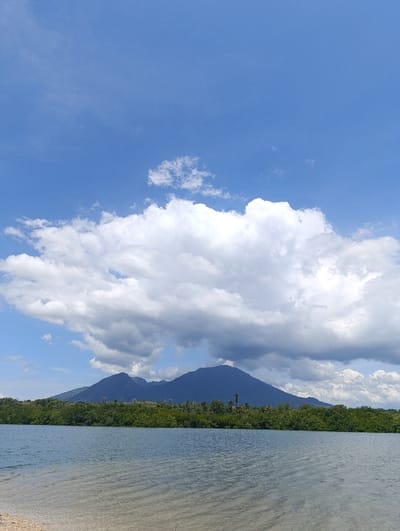 The image shows a calm body of water with a sandy beach in the foreground. The water is still, reflecting the blue sky and white clouds. There is a line of trees and bushes on the far side of the water. Behind the trees are mountains, dark green and blue, with a large white cloud hovering over them. The sky is a clear blue with scattered white clouds. The location is Kecamatan Banyuputih, Indonesia, at latitude: -7.754, longitude: 114.366. There are no people present.
