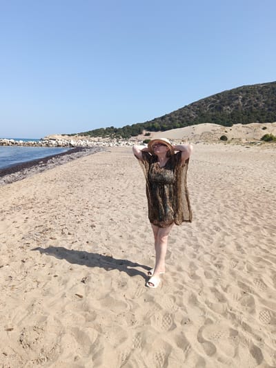 A woman wearing a patterned cover-up, a straw hat, and sunglasses stands on a sandy beach. Her hands are clasped behind her head as she looks up at the sky. She is facing forward, with her right foot slightly ahead of her left. The beach is covered in light brown sand, with scattered debris and footprints. In the background, there is a rocky shoreline, the sea, and a green, tree-covered hill. The sky is clear and blue. The location is identified as Municipal Unit of Larissos, Greece.