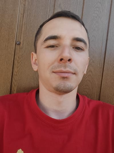 A close-up, eye-level, front-facing shot shows a young man with short, dark hair, a receding hairline, and light brown eyes. He has a thin mustache and a small goatee, and his skin is fair with some stubble. He is wearing a red t-shirt and a silver chain necklace. The background is a wooden wall with vertical planks. The lighting is natural and appears to be from the sun. The image was taken outdoors in Shkurupiivka, Ukraine.