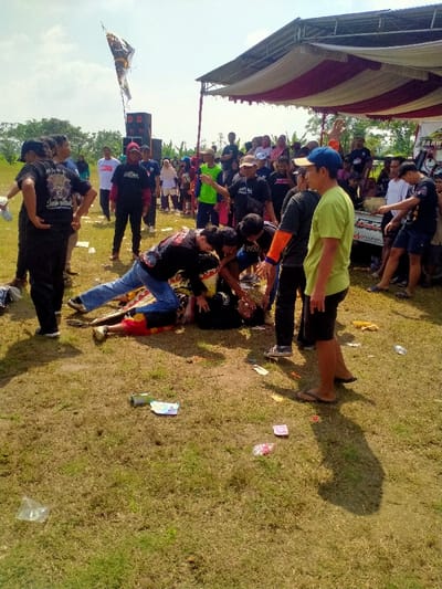 The image depicts an outdoor public event in Celep, Indonesia, under a clear sky with scattered clouds. The foreground and center show a cluster of individuals on a dry, grassy field littered with plastic wrappers and debris.
Central to the scene is a male individual lying supine on the ground, wearing a black shirt and red and yellow patterned pants. His face is upturned, with eyes closed or rolled back and mouth open, indicating an altered or distressed state. Four other men are actively engaged with him: one man in a black graphic t-shirt and blue jeans kneels at the head of the prone individual, with his hands positioned near the face/upper body. Another man in a black graphic t-shirt leans over, while a third in a black shirt with orange sleeves also attends. These individuals appear to be either assisting or ritually interacting with the person on the ground.
To the immediate left, a man in a black graphic t-shirt stands with his hands on his hips, observing. To the right, a man in a lime green t-shirt, blue baseball cap, black shorts, and flip-flops stands, partially turned away from the camera, also observing the central interaction.
In the mid-ground and background, a crowd of approximately 30-40 people, consisting of men, women (some wearing hijabs), and children, are gathered. Many are positioned under a large, elevated canopy structure featuring red and white striped fabric. To the left of this canopy, two large black audio speaker stacks are visible, with a pole extending upwards from which a dark banner or flag, possibly depicting an animal head, is suspended. The front section of a white or light-colored pickup truck, bearing partial text "SAHW," is visible under the right side of the canopy. The distant background shows sparse trees and vegetation under the bright sky.
