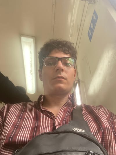 A male individual is depicted in a close-up selfie taken inside what appears to be a public transport vehicle, likely a train, in the Partido de Lanús, Argentina. The subject, with short, curly dark hair and dark-framed eyeglasses, wears a red and white vertical striped button-up shirt and a black sling bag across his chest with a circular silver emblem. He maintains a neutral expression, looking directly into the camera. The background consists of light-colored interior walls and ceiling. A rectangular fluorescent light fixture is visible on the upper left. On the upper right wall, a blue and white sign is affixed, featuring stylized male and female figures, indicative of a restroom, with partially visible Spanish text below them. A dark, indeterminate object or limb is partially visible behind the subject's left shoulder.