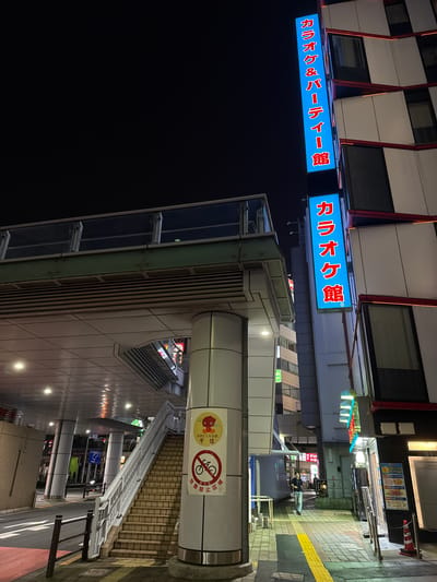 This is a nighttime, low-angle shot of a street scene in Adachi, Japan. A pedestrian walkway runs alongside a building with a prominent vertical blue sign that displays Japanese text in red, reading "カラオケ & パーティー館" (Karaoke & Party Hall) and "カラオケ館" (Karaoke Hall). Adjacent to the walkway, a set of tiled stairs ascends, bordered by a white railing and a cylindrical pillar. Attached to the pillar is a circular yellow sign with red text and graphics, depicting a bicycle with a red circle and line through it, and another graphic that appears to be a stylized red character. Further down the walkway, a person walks away from the camera. The street is paved with yellow and gray tiles. Buildings on either side of the walkway are illuminated.