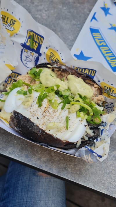 A close-up view displays a large, split baked potato served in a white cardboard tray, lined with 'Potato Star' branded paper, positioned on a light gray, textured surface. The potato has a dark skin and is filled with mashed potato, topped with two halves of a hard-boiled egg. Abundant chopped green onions and a sprinkle of black pepper cover the egg. A light green, creamy sauce is generously drizzled across the top, and other visible fillings include a flaked substance, possibly tuna, and dark, irregular pieces. The 'Potato Star' branding on the paper wrapper features a cartoon face in a top hat, text reading 'FULL OF goodieS,' and Hebrew text 'פוטטו סטאר' (Potato Star). In the lower left background, a person's lower leg, clad in ripped blue jeans, is visible, suggesting an individual is seated at the surface. The scene is located in Kiryat Gat, Israel.