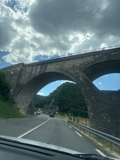 The image captures a low-angle view of a large stone viaduct, likely a railway bridge, with its arches framing a lush, green mountainside and a bright blue sky interspersed with white, fluffy clouds. The stone construction of the viaduct is detailed, showing individual blocks and the curved architecture. A modern metal guardrail runs alongside a paved road visible to the right of the frame, indicating a route passing beneath or near the viaduct. Several cars are seen on the road, suggesting ongoing traffic. The overall scene is bathed in daylight, with the sun creating bright patches on the clouds and illuminating the landscape. The orientation of the image is rotated, making it appear as if the viewer is looking upwards from ground level or from a vehicle driving along the road. The setting appears to be a natural, possibly mountainous area, given the dense vegetation on the slopes. No people are directly visible, but their presence is implied by the cars on the road. The time of day appears to be daytime, likely late morning or afternoon due to the bright sunlight and cloud cover. There is a speed limit sign visible on the side of the road with "60" indicating kilometers per hour. The location is identified as Hauts de Bienne, France.